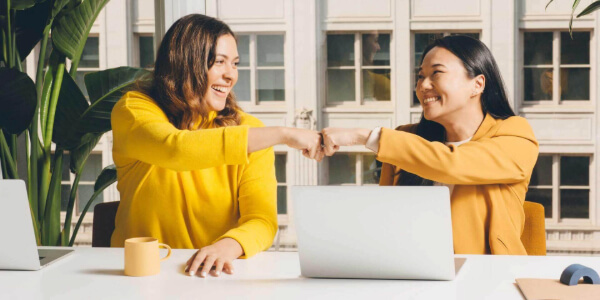 Dos mujeres sonrientes se dan un choque de puños en una oficina luminosa. Ambas visten ropa amarilla y están sentadas frente a un escritorio con computadoras portátiles y una taza de café. En el fondo, se pueden ver grandes ventanales que permiten la entrada de luz natural.