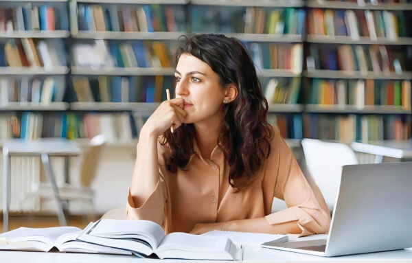 Una mujer sentada en una mesa de estudio, con libros abiertos frente a ella y una computadora portátil a su lado. Ella mira pensativa hacia un lado, con una mano en la boca, en un ambiente de biblioteca con estanterías llenas de libros al fondo.