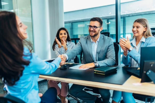 Un grupo de cuatro profesionales en una sala de reuniones. Una mujer con camisa azul está estrechando la mano de un hombre con gafas y traje gris, mientras los otros dos miembros del equipo aplauden y sonríen, mostrando una atmósfera de celebración y éxito.