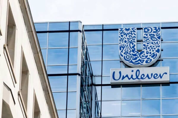 Fachada de un edificio con paneles de vidrio, destacando el logo de Unilever en la parte superior. El logo presenta un diseño artístico en azul, mientras que el nombre 'Unilever' está escrito en letras azules sobre un fondo blanco.